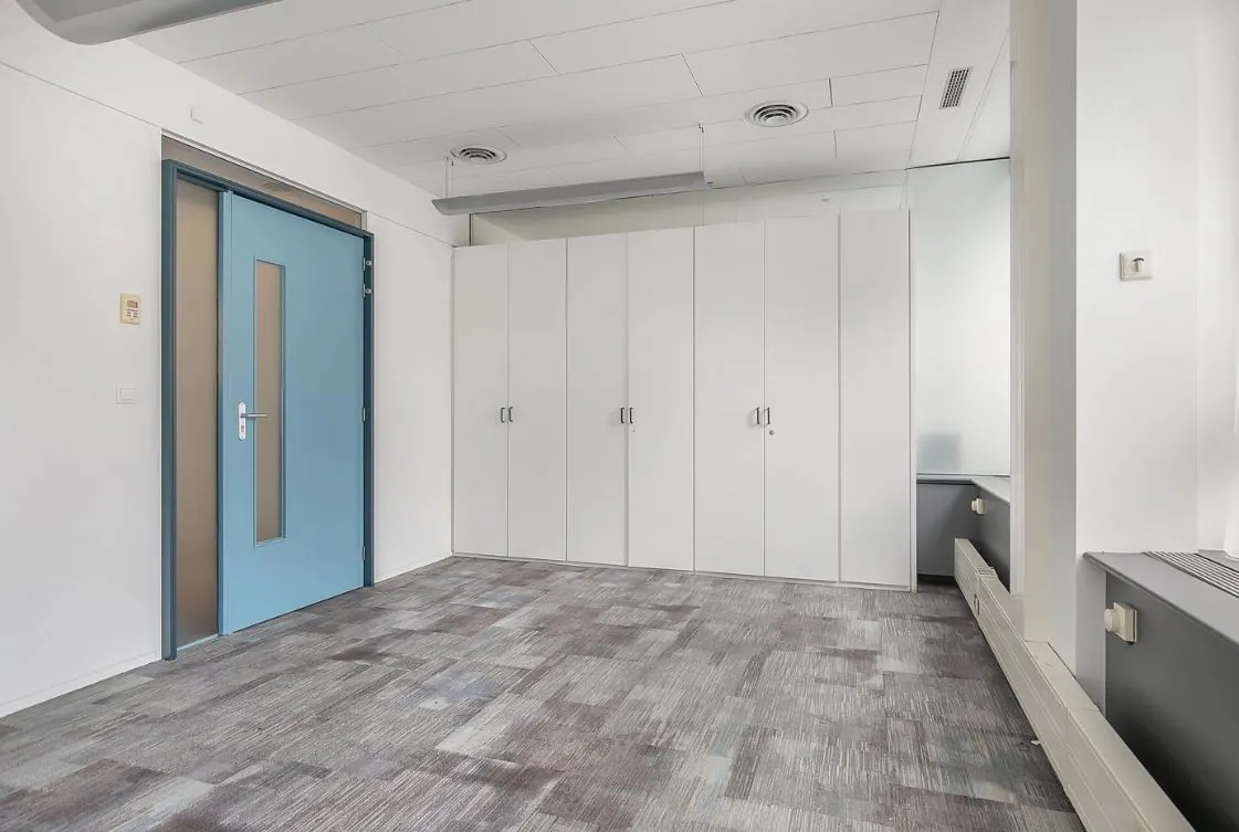 Leeg kantoor met grijze vloerbedekking, witte kastenwand en een blauwgrijze deur.