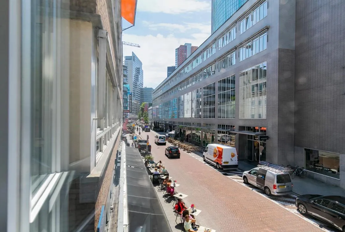 Straatbeeld van de Meent in Rotterdam met terrassen, voetgangers, auto's en moderne kantoorgebouwen op een zonnige dag.
