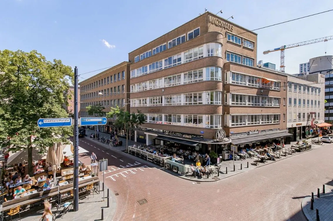 Straatbeeld met het Minervahuis in Rotterdam, waar mensen op terrassen zitten bij café-restaurant en wijnbar NRC.
