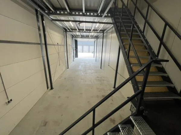 Interieur van een lege bedrijfsruimte aan de Aalsmeerderweg met metalen trap en deels open garagedeuren op de achtergrond.
