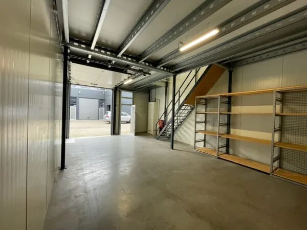 Interieur van een lege bedrijfsruimte aan de Aalsmeerderweg met metalen trap, open garagedeur en houten stellingen.