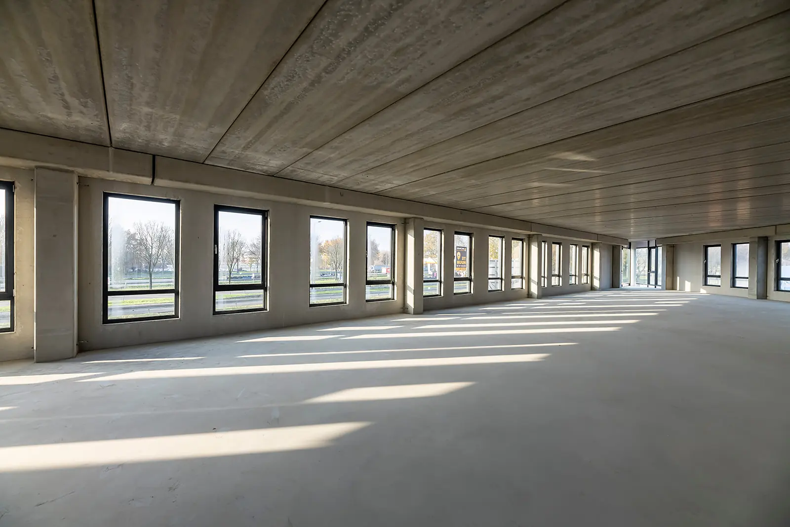 Leeg betonnen kantoorpand met grote ramen aan de Kringloopweg, waar zonlicht naar binnen valt.