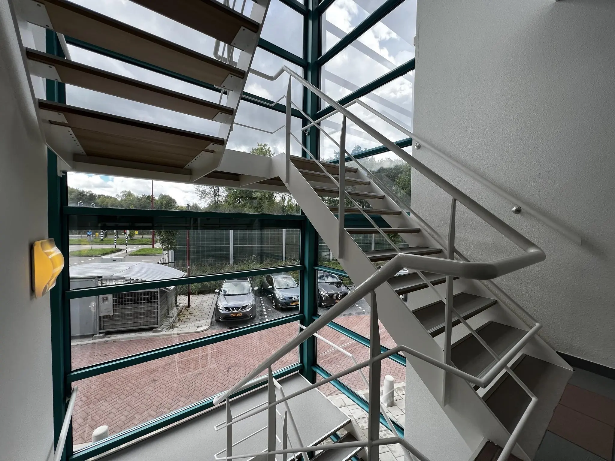 Trapportaal met metalen trap en grote ramen in het gebouw Zoutkamperschans, met uitzicht op geparkeerde auto's en een kruispunt buiten.