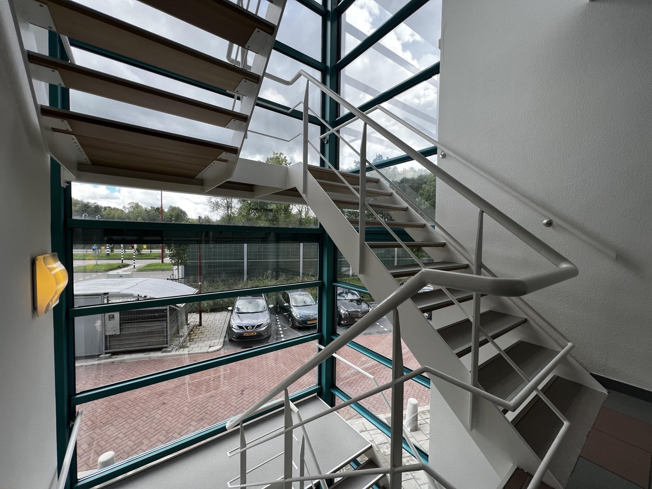 Het interieur van een moderne trap met een metalen leuning en grote ramen met uitzicht op geparkeerde auto's.