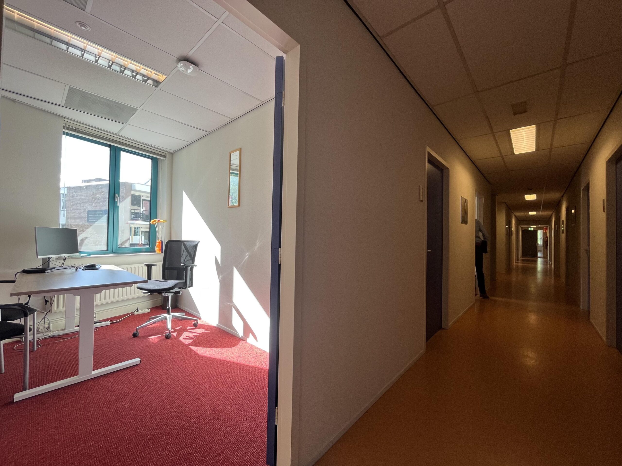 Een kantoor met een bureau en stoel kijkt uit op een lange gang met gesloten deuren.