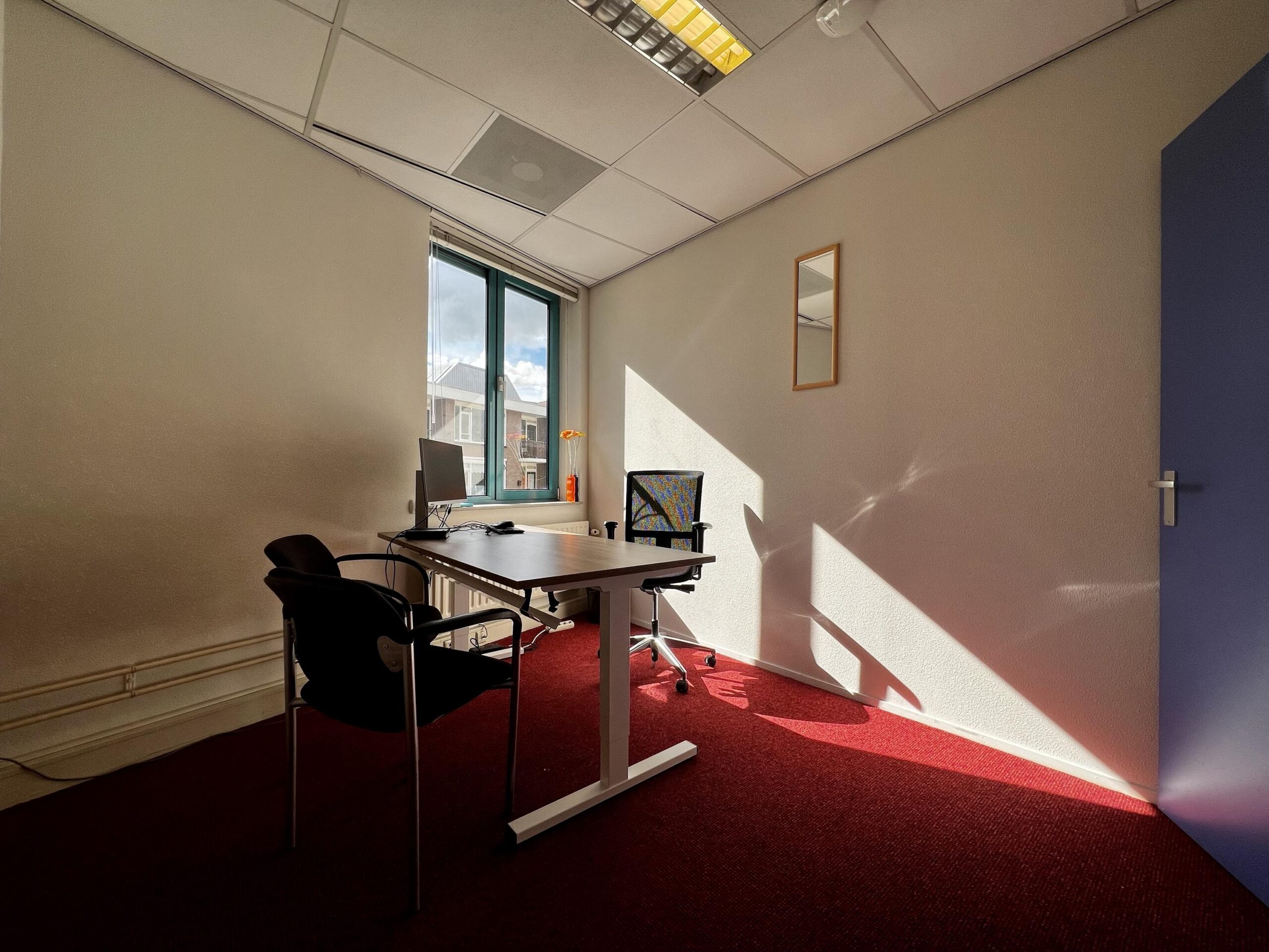 Een klein kantoor met een bureau, twee stoelen, een computer en een raam dat uitkijkt op een gebouw.