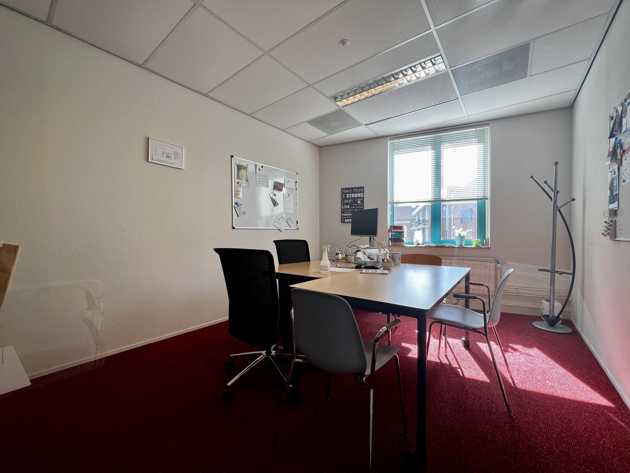 Een kleine vergaderruimte met een rood tapijt, een tafel met stoelen, en een raam dat zonlicht binnenlaat.