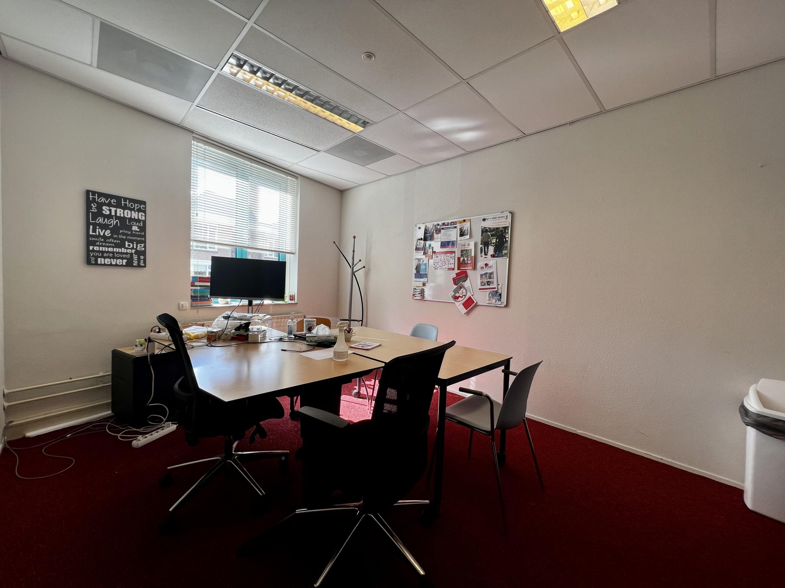 Een kantoorruimte met een tafel, stoelen, een computer en een prikbord met posters aan de muur.