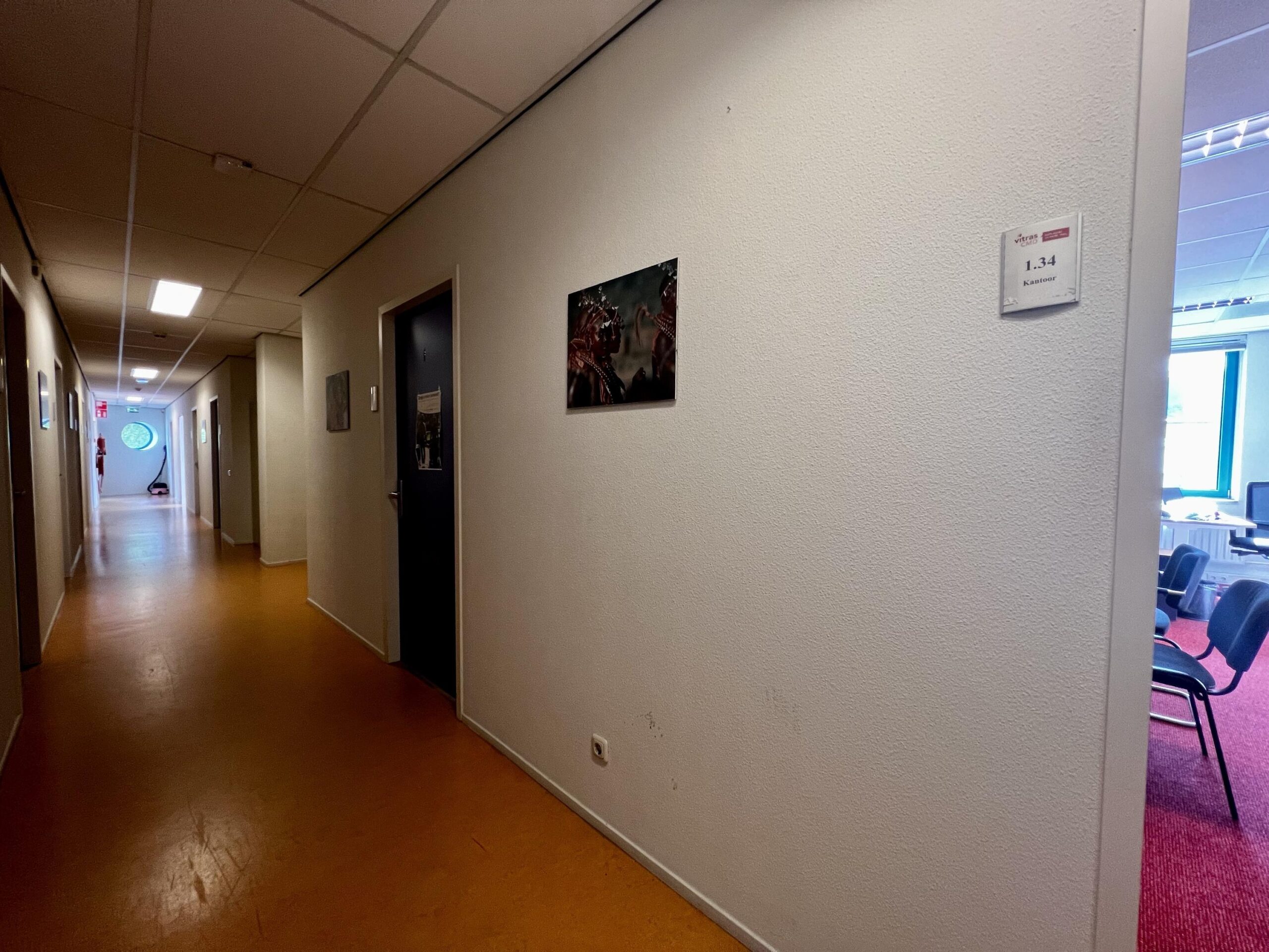 Een lange, smalle gang in een kantoor met links een brandblusser bij de nooduitgang en rechts een open deur naar een kantoorruimte.