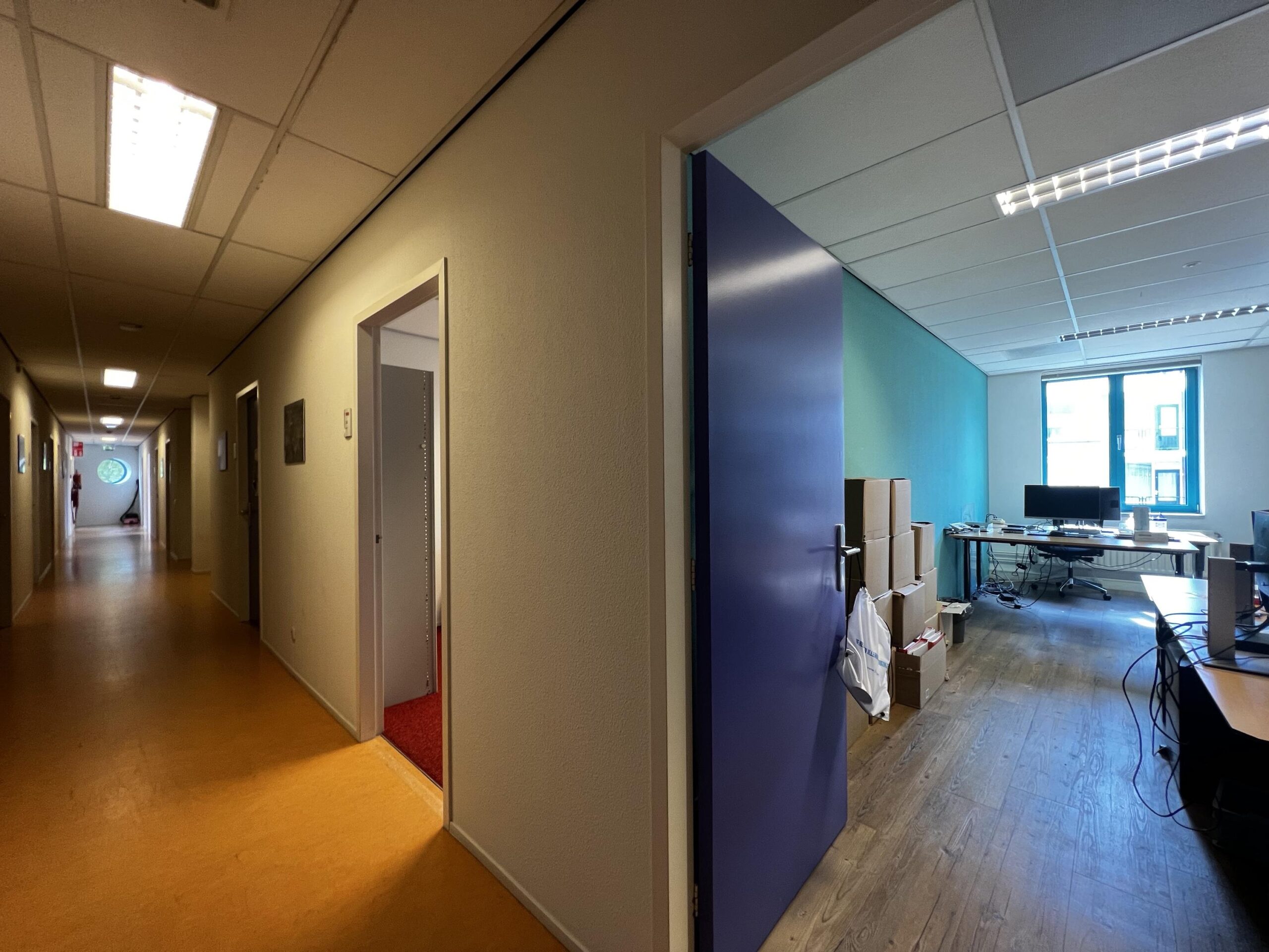 Een kantoor in een gang met open deur, gevuld met bureaus, dozen en kabels.