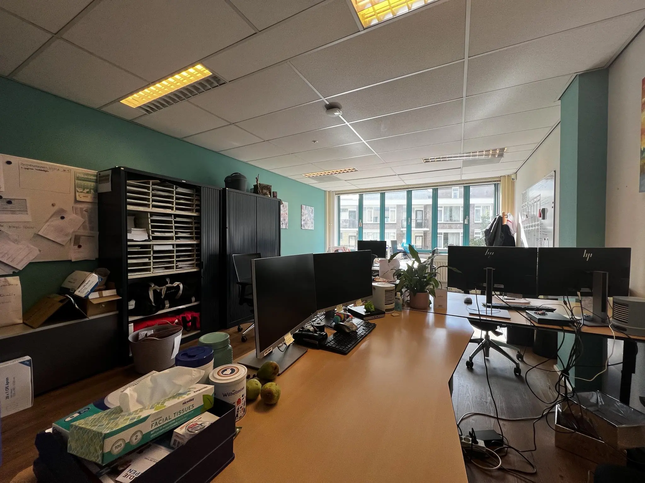Kantoorruimte aan de Zoutkamper Schans met bureaus, computerschermen, kasten en een groot raam met uitzicht op een appartementencomplex.