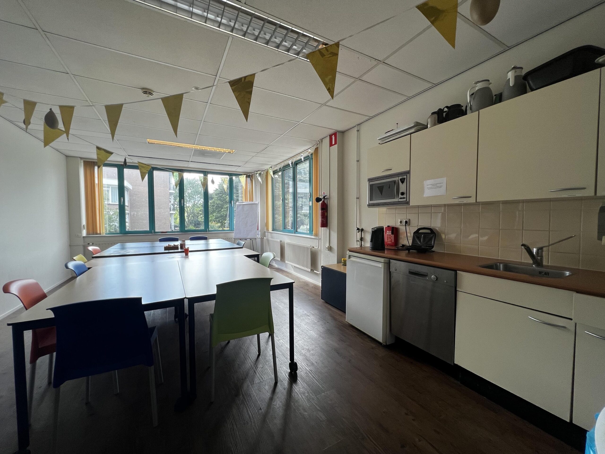 Een gemeenschappelijke ruimte met een keukenblok en meerdere tafels en kleurrijke stoelen.