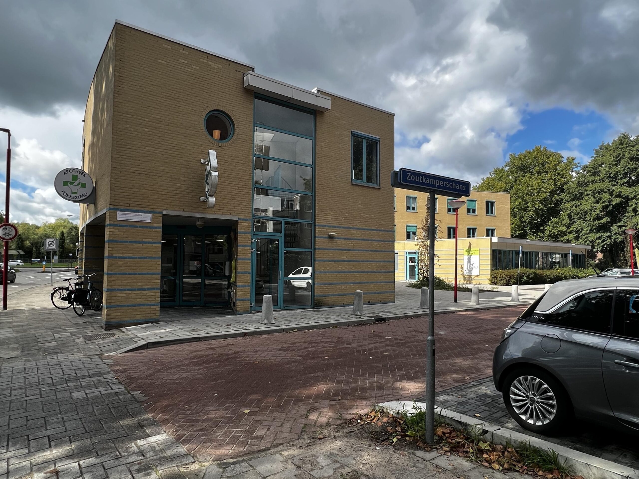 Een modern gebouw met het bord "Zoutkamperschans" en de apotheek "Vokesteeg" naast een straat met geparkeerde auto.