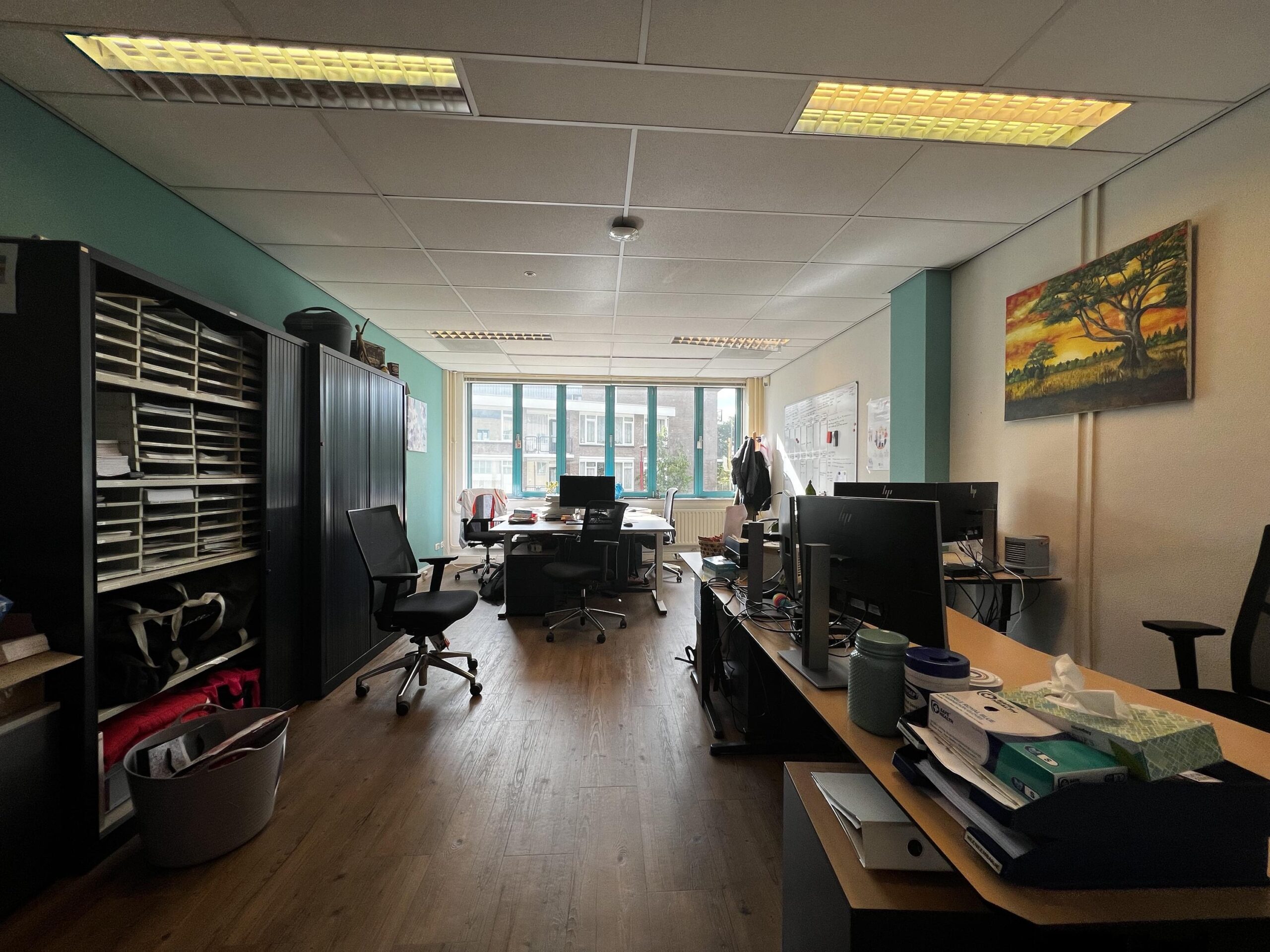 Een kantoorruimte met bureaus, stoelen, en computers, verlicht door plafondlampen, met een groot raam en schilderij aan de muur.