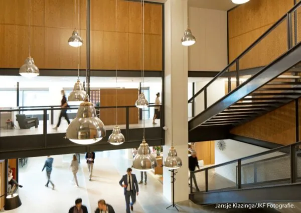 Moderne kantoorruimte in Hofplein met stalen trap, houten wanden en hanglampen, waar mensen rondlopen en werken.