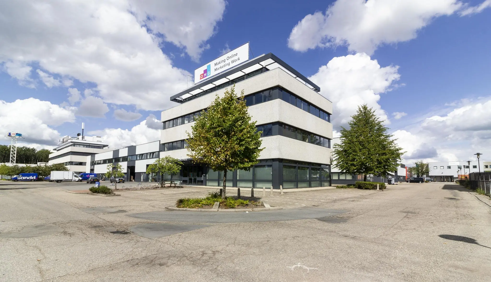 Kantoorpand van OMG aan de Zonnebaan in een bedrijventerrein met bomen en geparkeerde voertuigen.