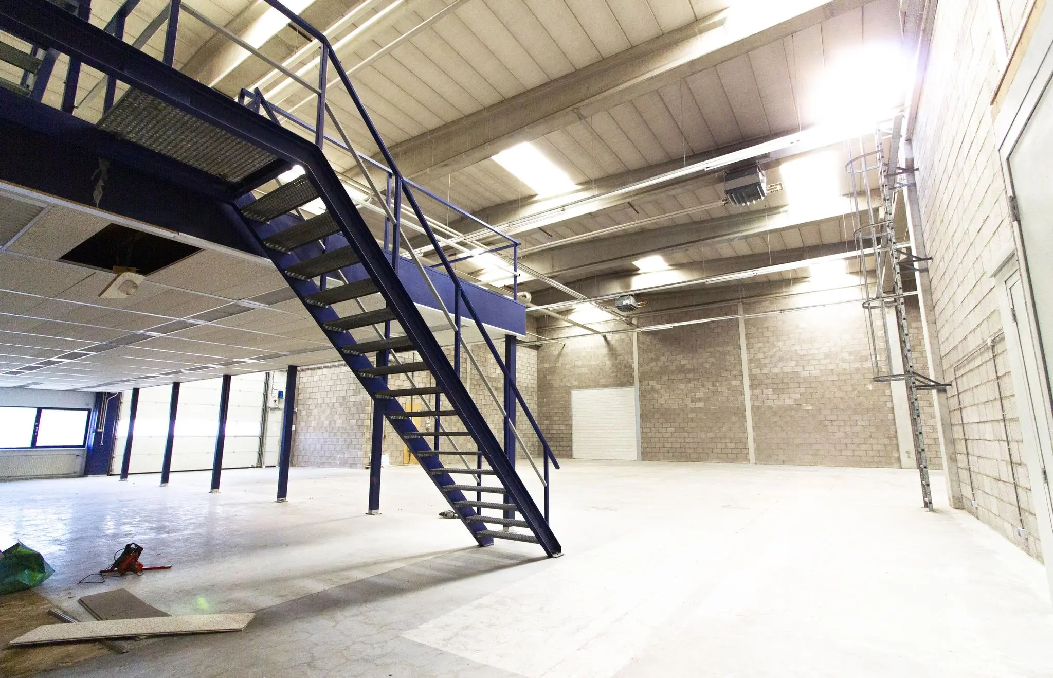 Ruime, lege bedrijfshal aan de Zonnebaan met metalen trap naar verdiepingsvloer.