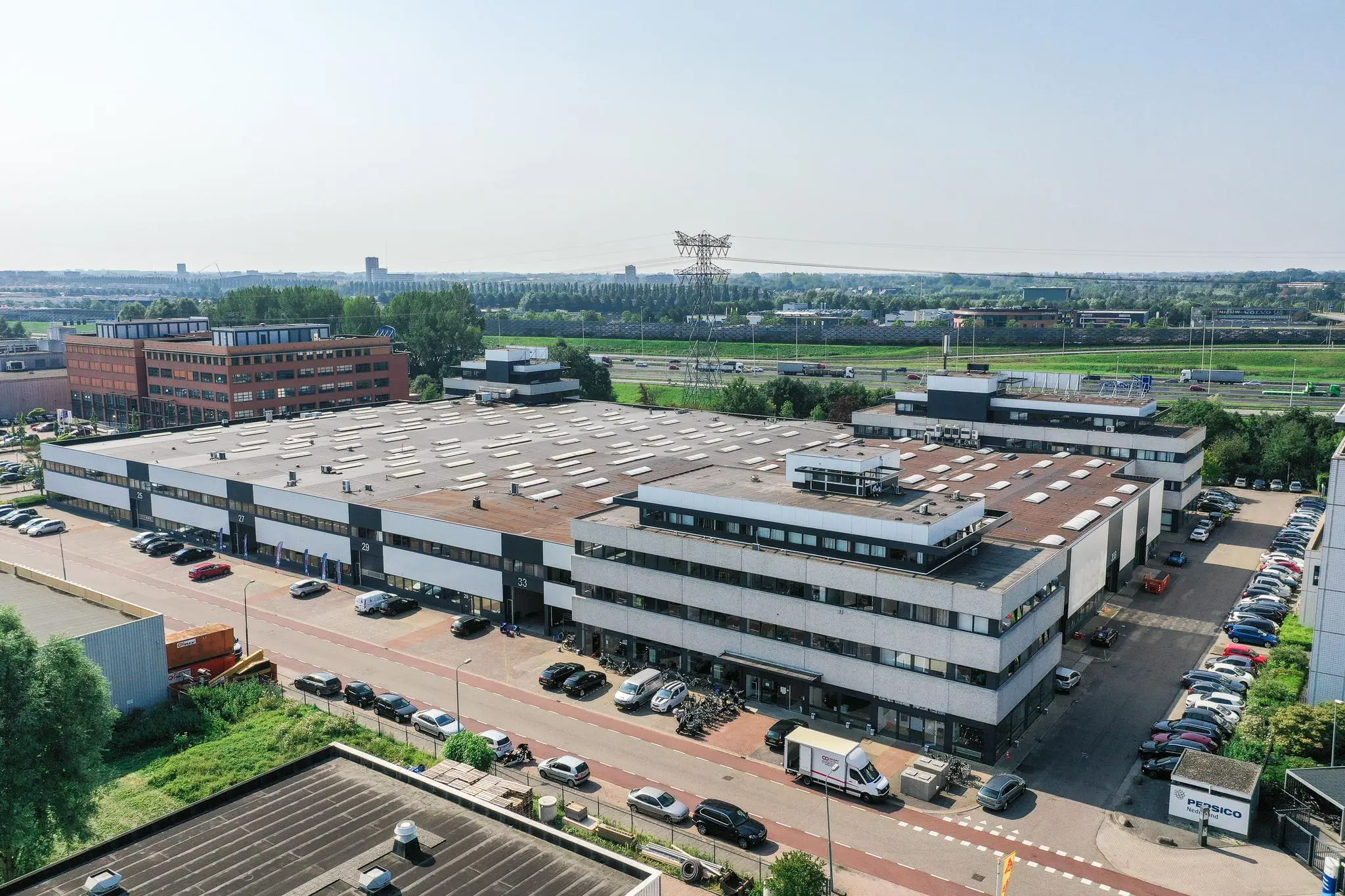 Luchtfoto van een bedrijventerrein aan de Zonnebaan met meerdere bedrijfsgebouwen, parkeerplaatsen en de snelweg op de achtergrond.