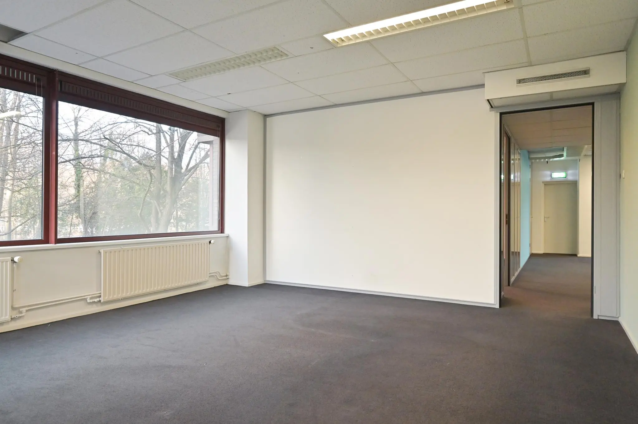 Lichte, lege kantoorruimte aan de Bezuidenhoutseweg met groot raam en uitzicht op bomen.