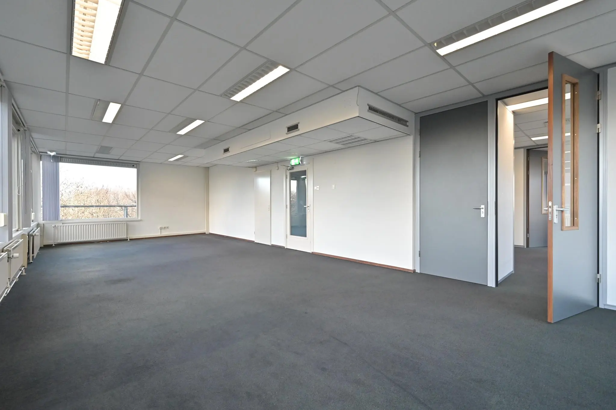 Lichte, lege kantoorruimte aan de Bezuidenhoutseweg met systeemplafond, grijze vloerbedekking en meerdere deuren.