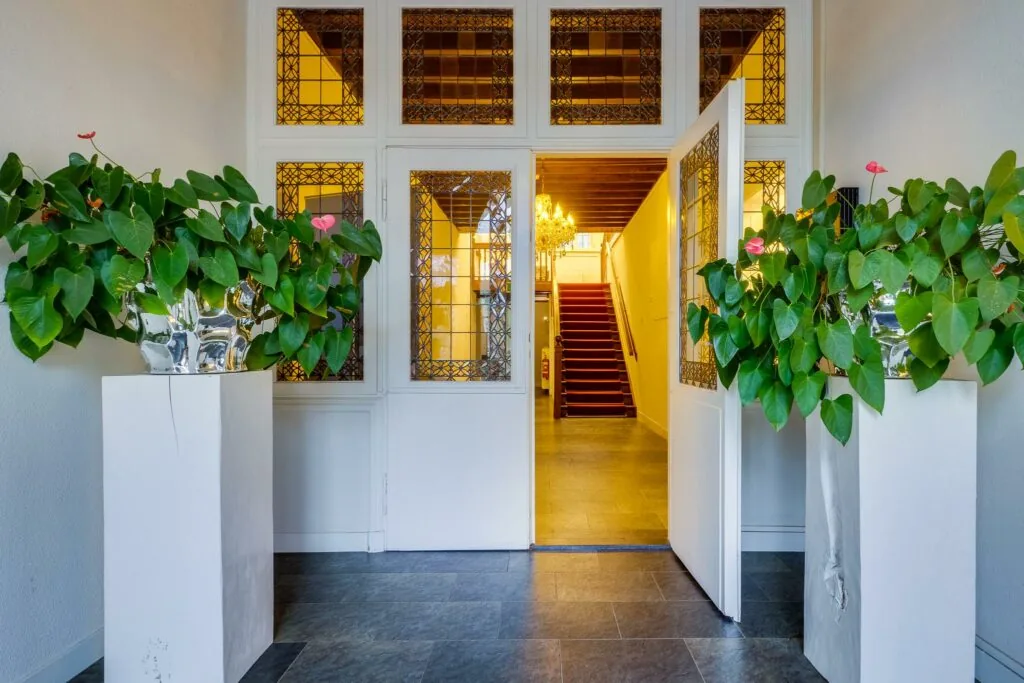 Ingang van een stijlvol pand aan de Amersfoortsestraat met glas-in-looddeuren, rode trap en grote groene kamerplanten in zilverkleurige potten.