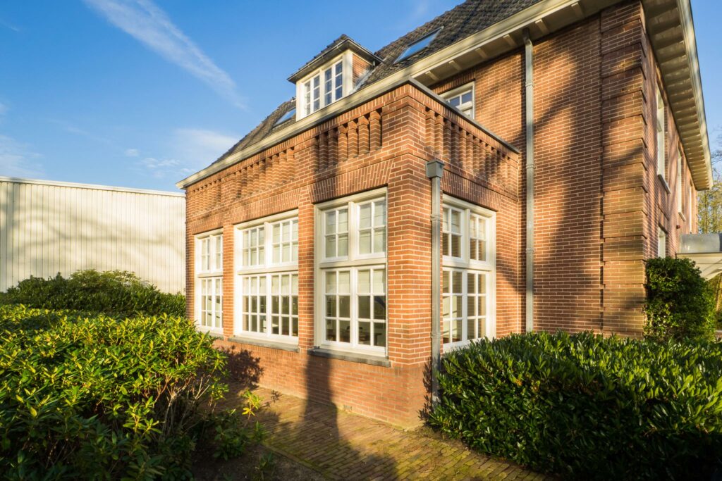 Rode bakstenen hoekpand aan de Amersfoortsestraat met witte raamkozijnen en groene struiken op een zonnige dag.