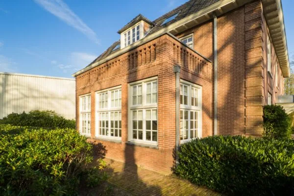Rode bakstenen hoekpand aan de Amersfoortsestraat met witte raamkozijnen en groene struiken op een zonnige dag.