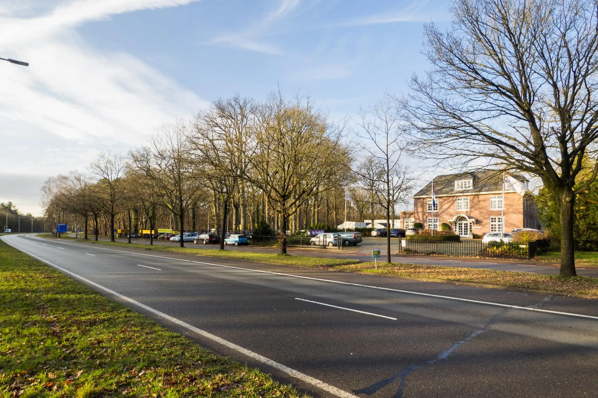 Brede straat met bomen langs de Amersfoortsestraat, naast een klassiek bakstenen kantoorpand en parkeerplaats.