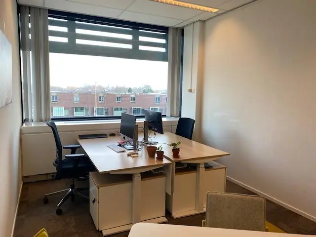 Kleine kantoorruimte aan de Oudlaan met twee bureaus, computerschermen en uitzicht op een woonwijk.