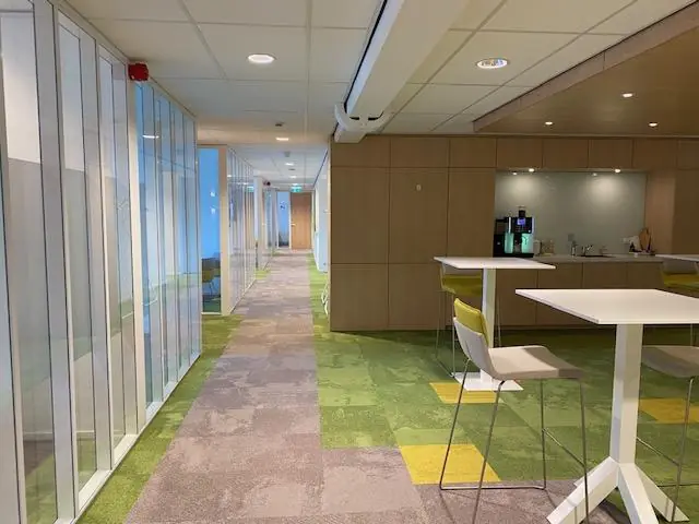 Moderne kantoorruimte aan de Oudlaan met glazen wanden, een groen tapijt en een koffiehoek met hoge tafels en stoelen.