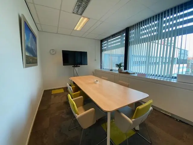 Vergaderruimte aan de Oudlaan met een lange tafel, zes stoelen, een scherm aan de muur en grote ramen met jaloezieën.
