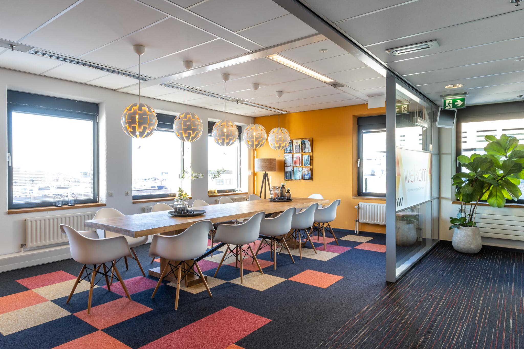 Vergaderruimte met lange houten tafel, moderne witte stoelen, kleurrijk tapijt en grote ramen aan de Oudlaan.