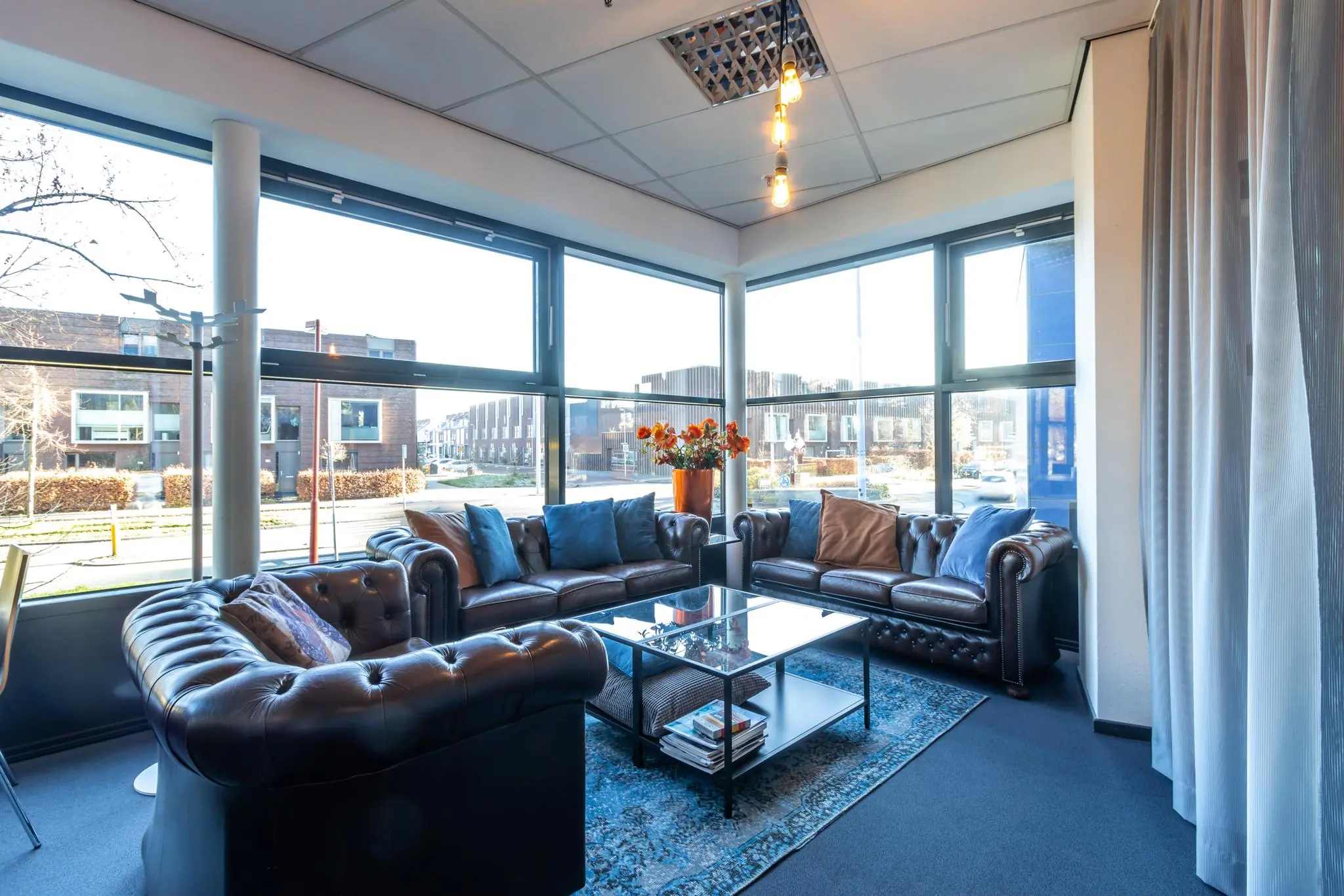 Zonnige wachtruimte aan de Oudlaan met leren chesterfieldbanken, glazen salontafel en grote ramen met uitzicht op moderne woongebouwen.