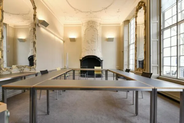 Vergaderruimte aan het Rapenburg met U-vormige tafelopstelling, hoge ramen, sierlijke spiegels en een rijk gedecoreerde schouw.