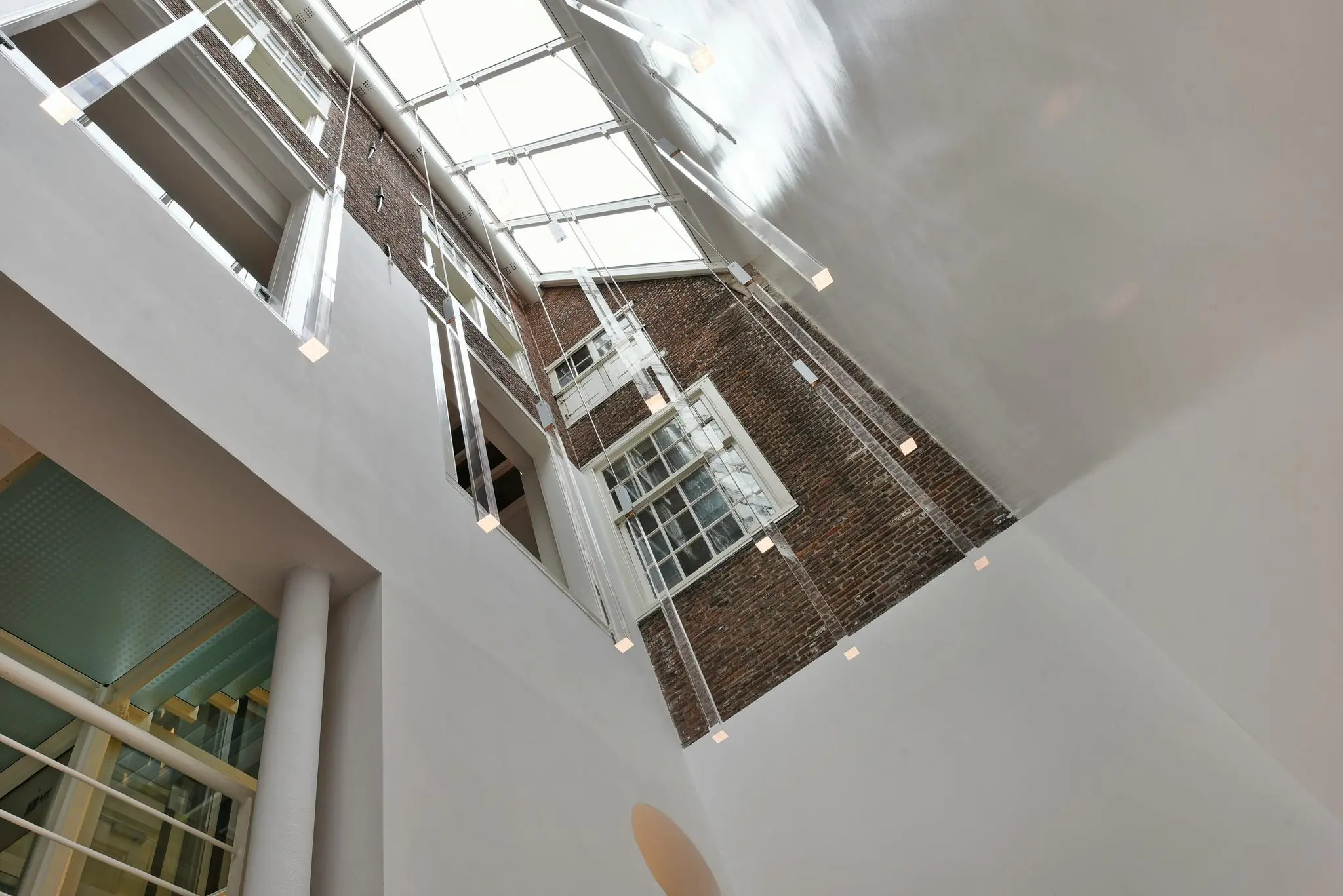 Doorkijk naar een glazen dak in het Rapenburggebouw met moderne hanglampen en zicht op een historische bakstenen gevel.