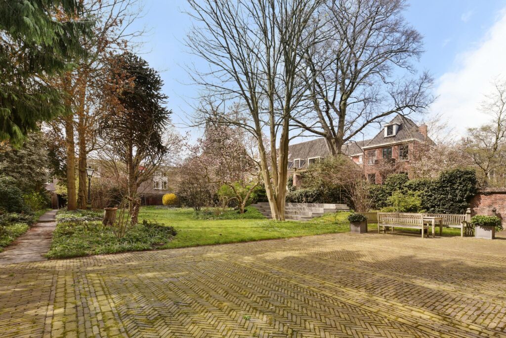 Grote, groene stadstuin aan het Rapenburg met oude bomen, zitbanken en een monumentaal pand op de achtergrond.