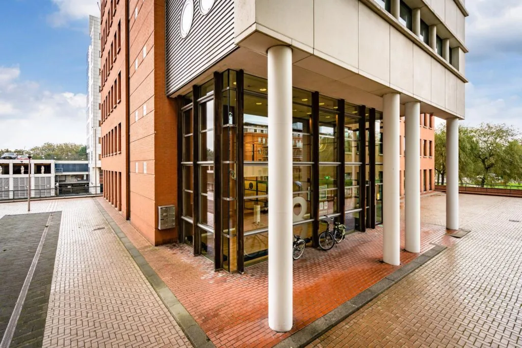 Ingang van een modern kantoorgebouw aan het Maanplein met hoge glazen ramen, witte zuilen en twee fietsen ervoor.