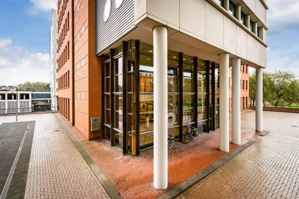 Ingang van een modern kantoorgebouw aan het Maanplein met hoge glazen ramen, witte zuilen en twee fietsen ervoor.