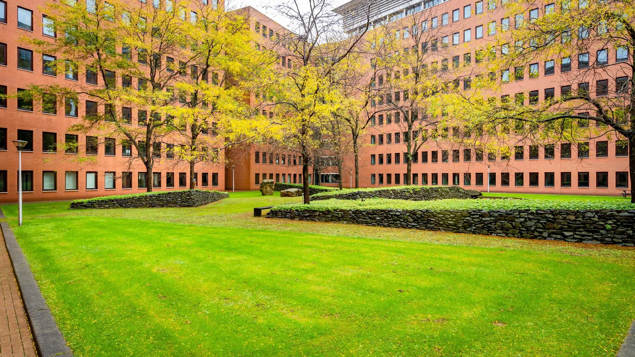 Binnenplein van het Maanplein in Den Haag met groene grasvelden, herfstbomen en een omliggend rood bakstenen kantoorgebouw.