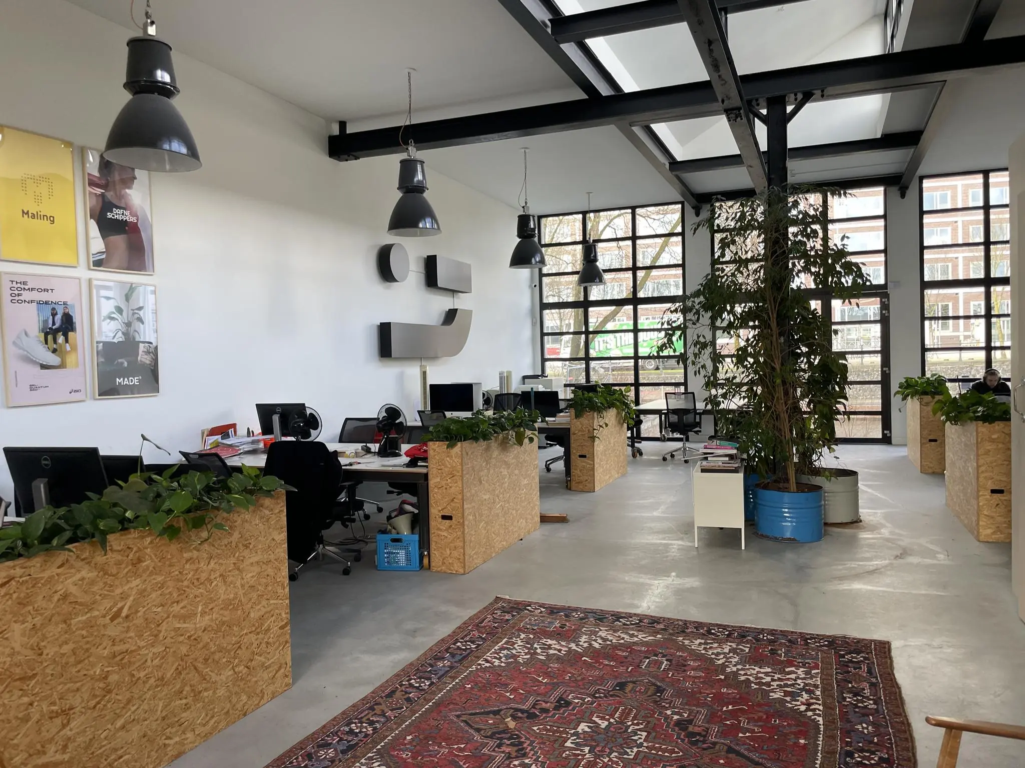 Moderne open kantoorruimte met houten bureaus, veel planten, grote ramen en industriële verlichting.