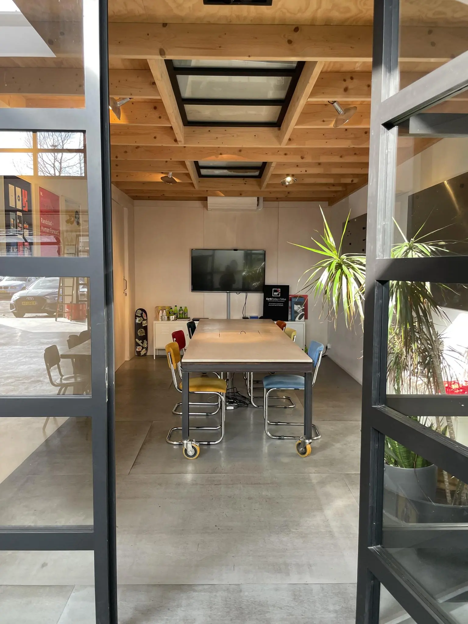 Vergaderruimte met houten plafond, glazen dakramen, lange tafel met gekleurde stoelen en een televisie aan de muur.
