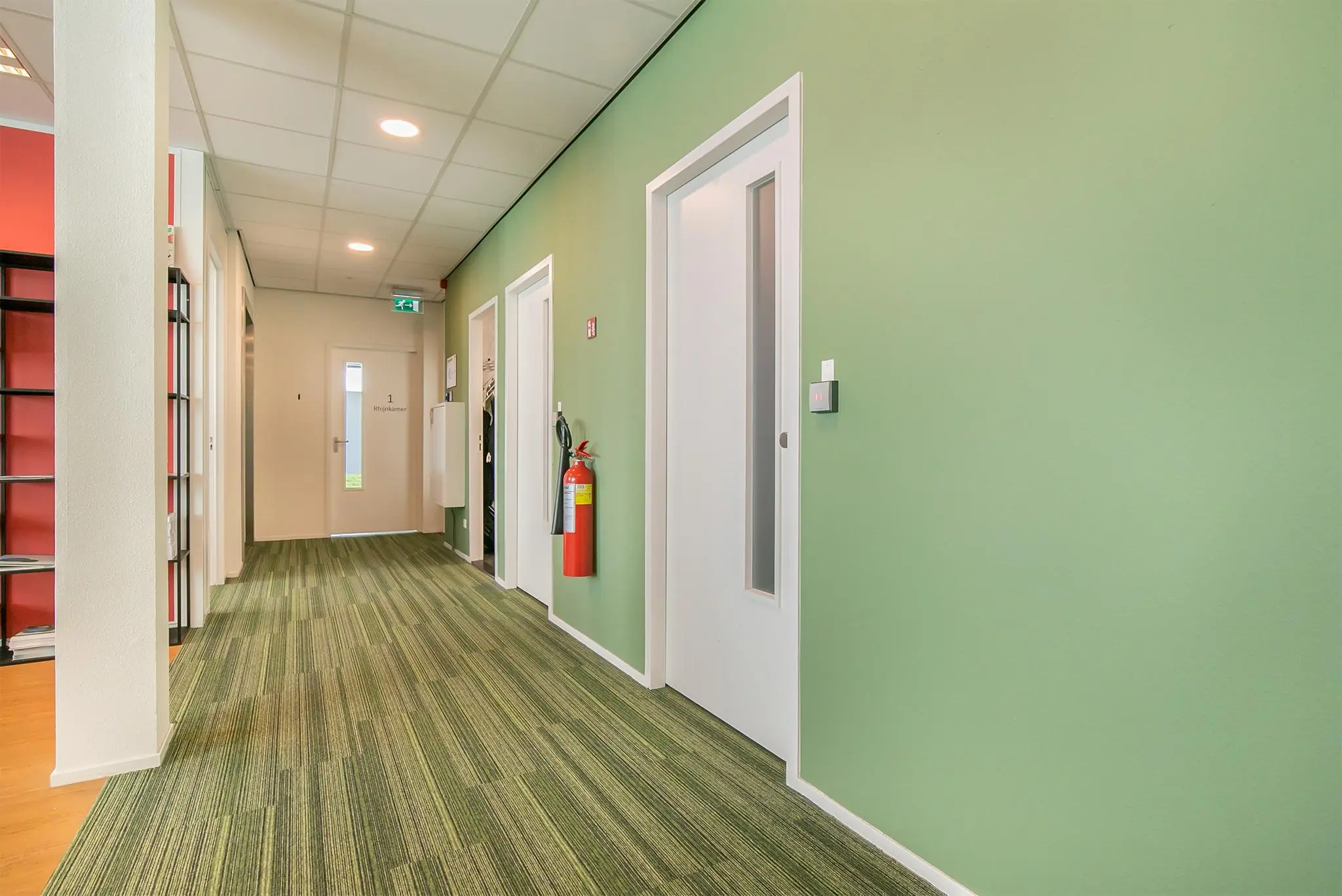 Gang met groene muren en vloerbedekking, brandblusser aan de muur en deur met opschrift "Rhijnkamer 1".