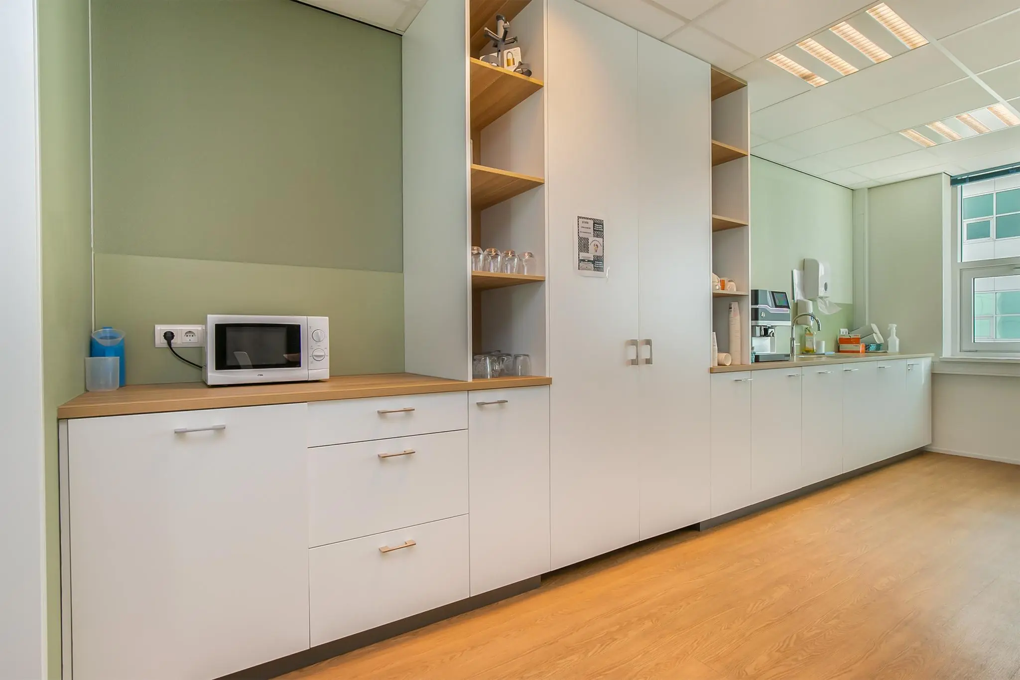 Moderne pantry in een kantoor met witte kasten, een magnetron, koffieautomaat en houten vloer.