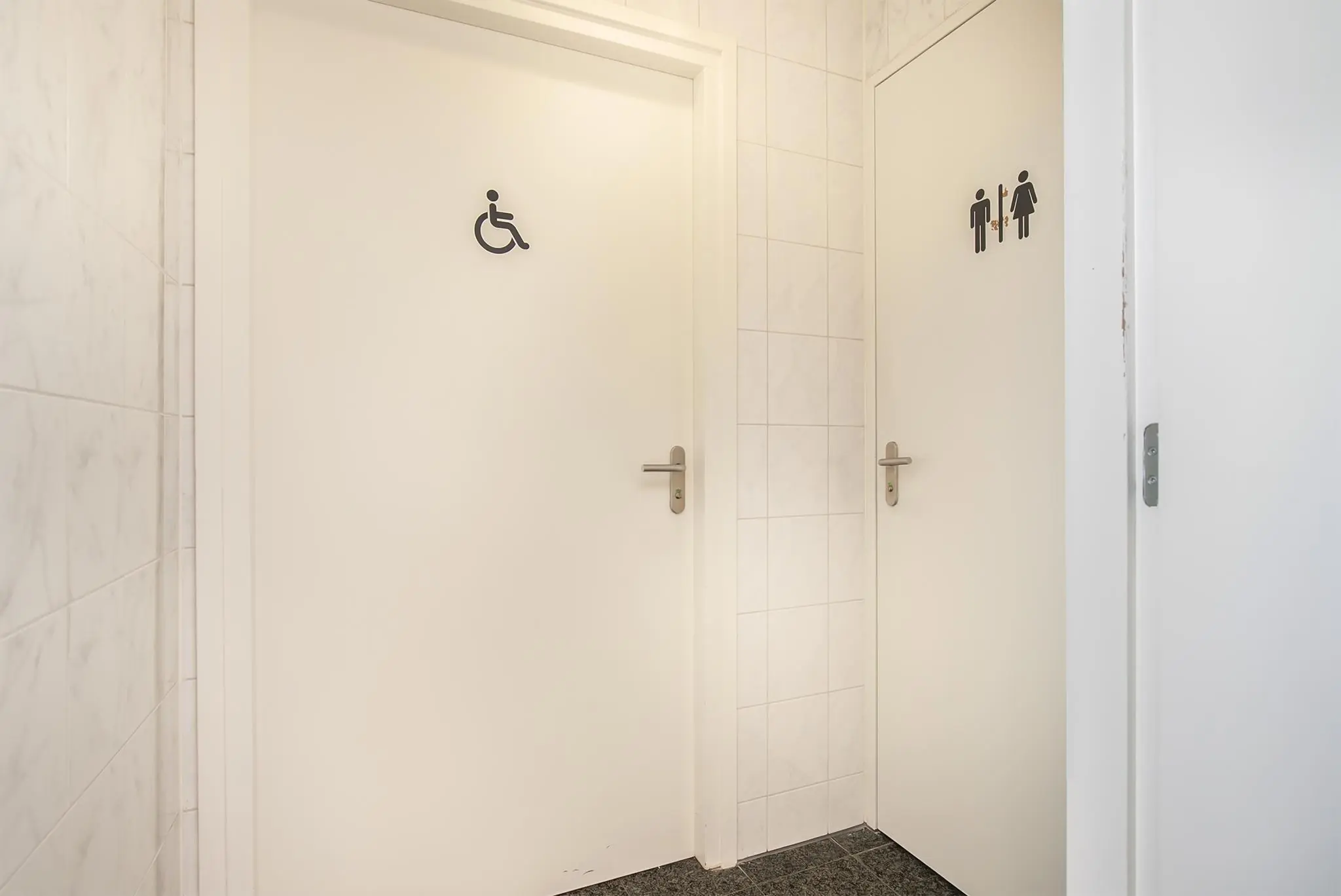 Twee witte toiletten met pictogrammen voor rolstoelgebruikers en een uniseks toilet in een betegelde gang.