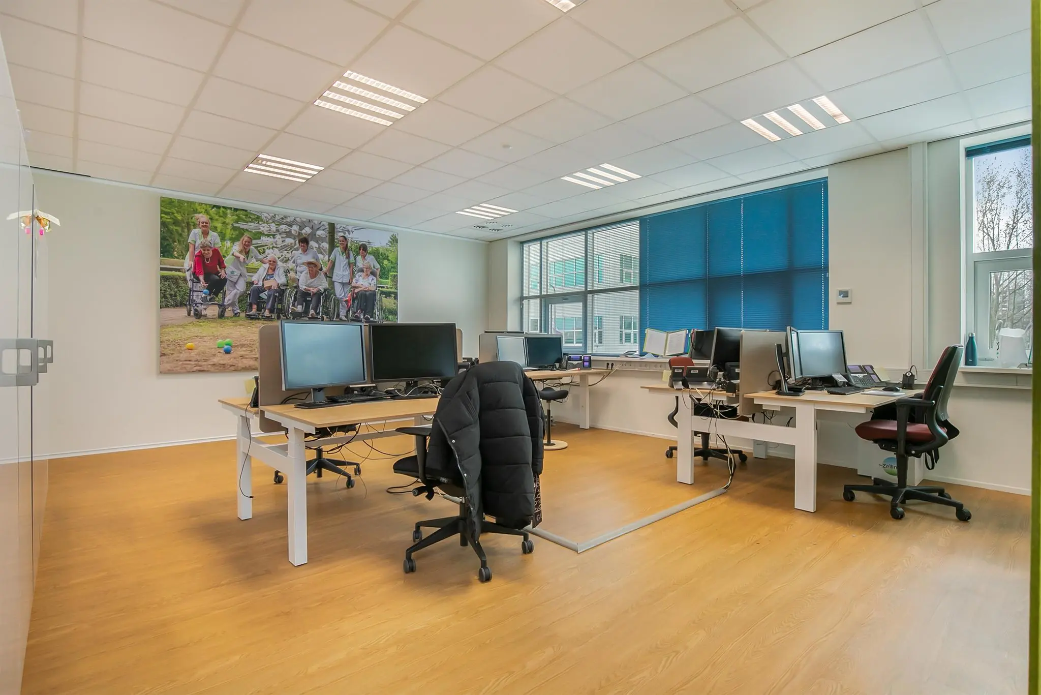 Modern kantoor met meerdere werkplekken en een grote foto aan de muur van een groep mensen met rolstoelen in een park.