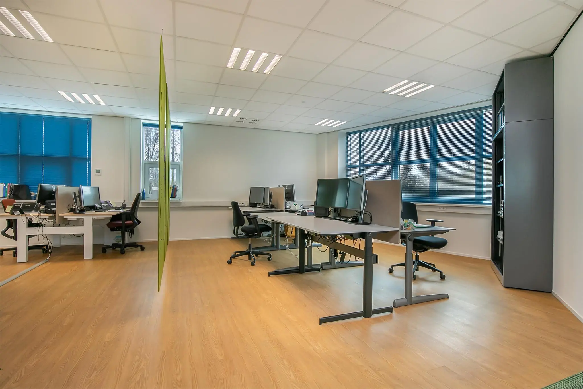 Modern en licht kantoor met meerdere werkplekken, computers en grote ramen met blauwe jaloezieën aan het Rhijnspoor.