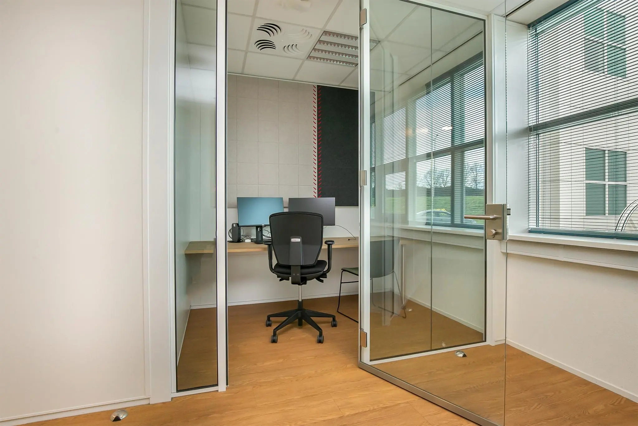 Kleine moderne kantoorruimte met glazen wanden, een bureau met twee schermen en een bureaustoel.