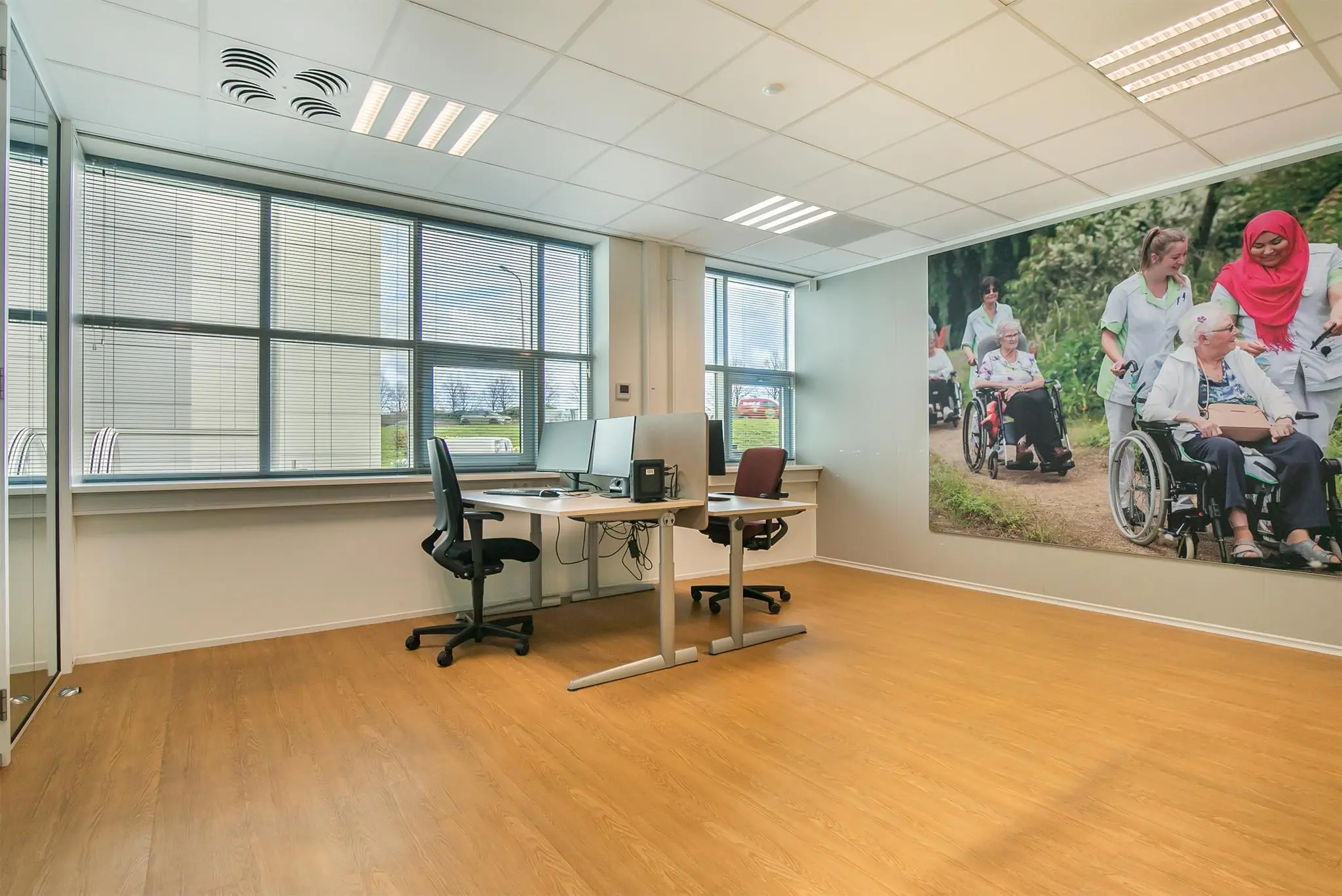 Modern kantoor met twee bureaus en een grote wandfoto van zorgverleners met ouderen in rolstoelen bij Rhijnspoor.