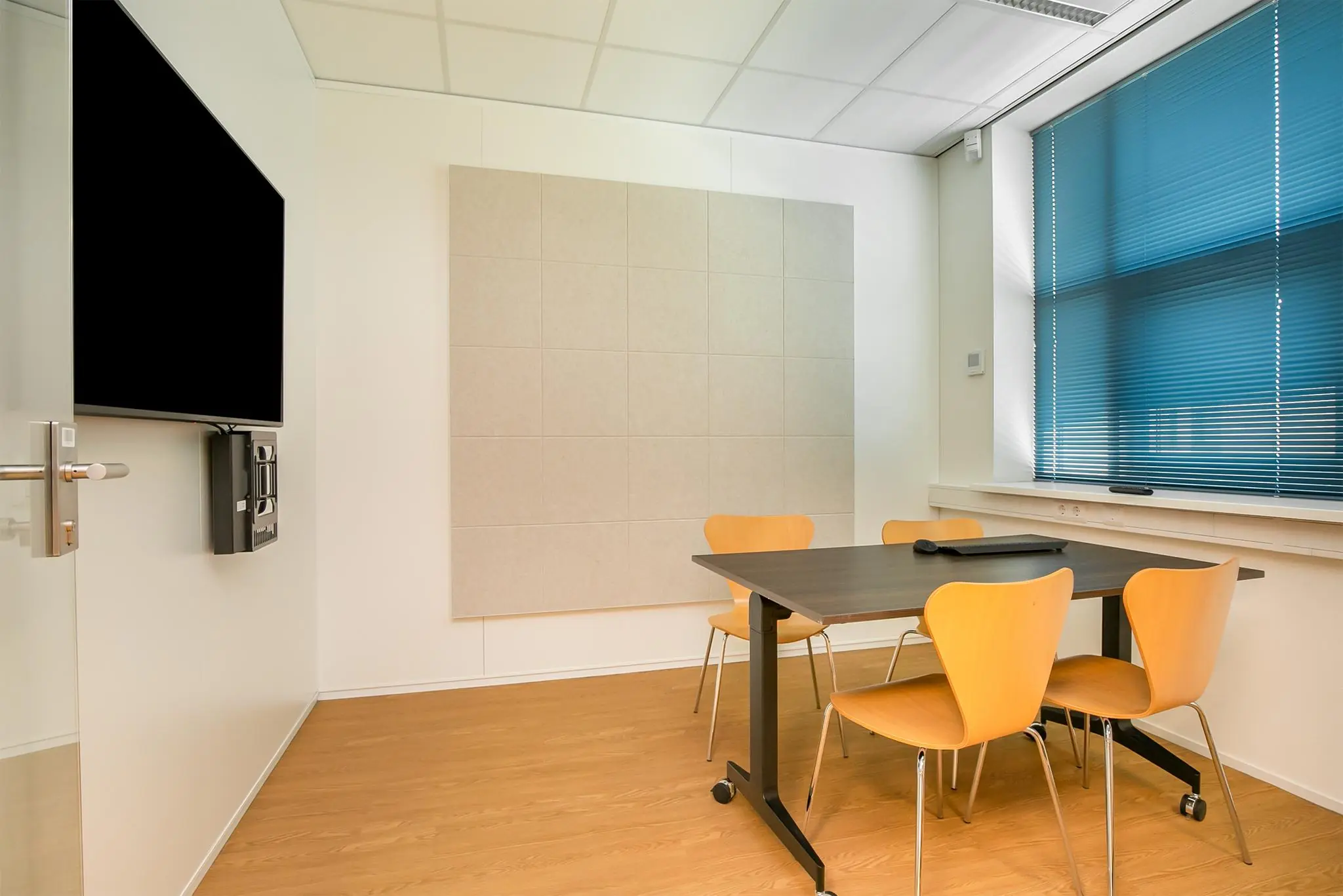 Kleine vergaderruimte met vier houten stoelen, een zwarte tafel, een scherm aan de muur en blauwe jaloezieën voor het raam.