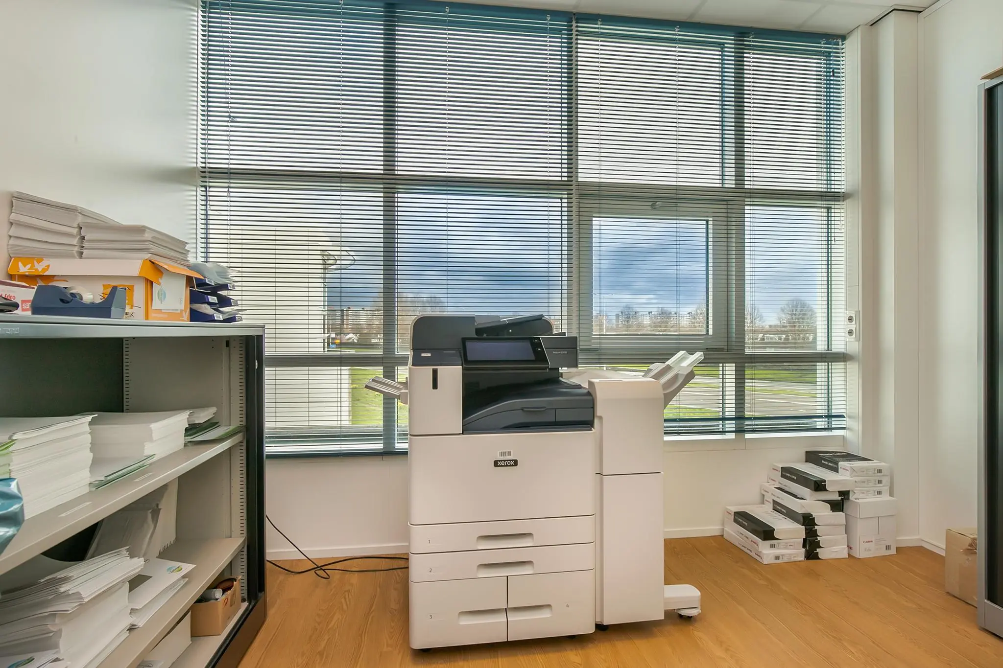 Multifunctionele Xerox-printer in een kantoorruimte met houten vloer en grote ramen met jaloezieën.